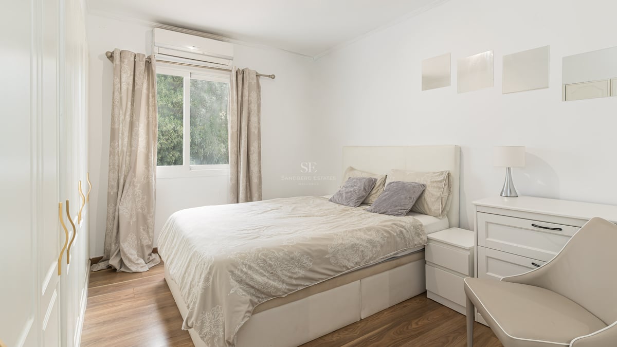 Chambre lumineuse avec lit double, meubles blancs, parquet et fenêtre donnant sur des arbres.