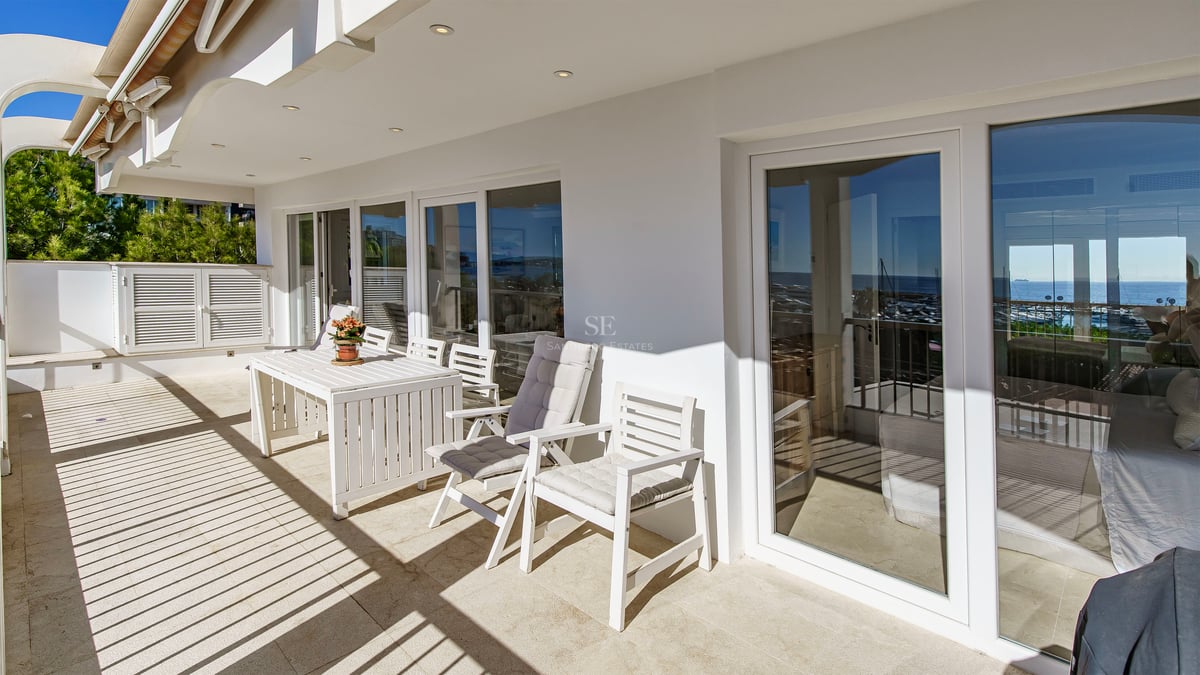 Sunlit terrace with white outdoor dining set and large glass sliding doors reflecting the coastline.