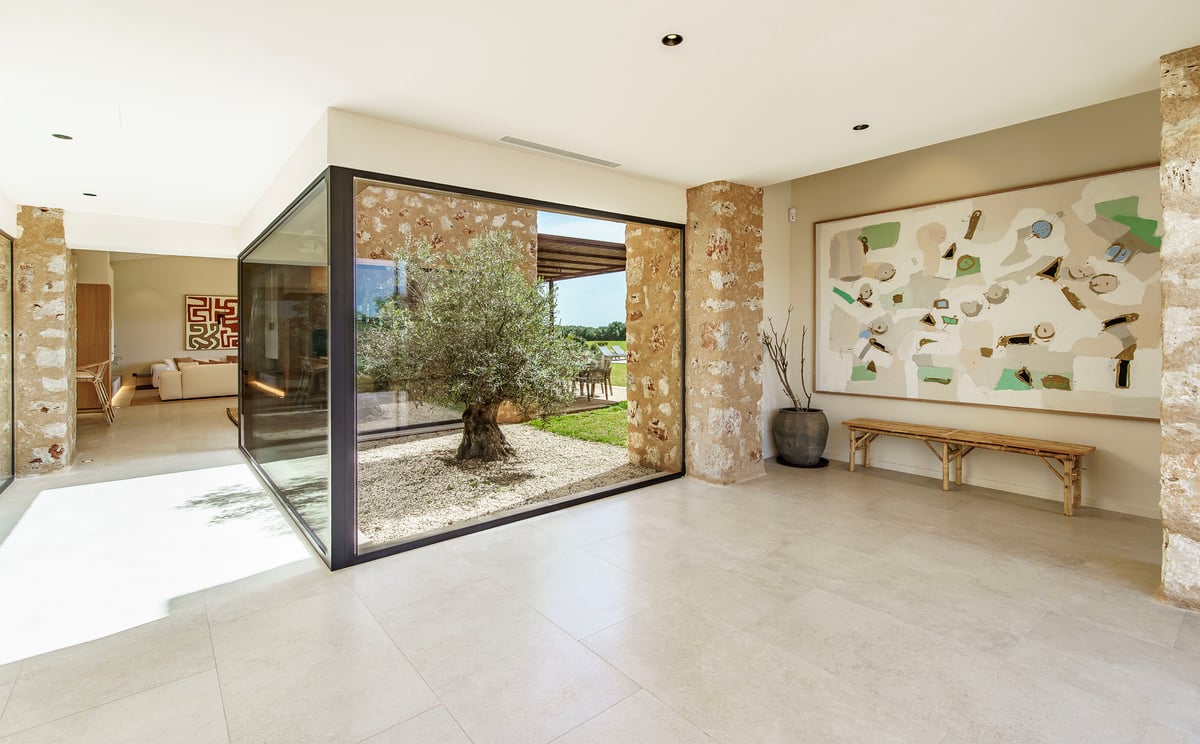 Moderner Flur mit bodentiefen Glasfenstern, die einen Innenhof mit einem Olivenbaum und Natursteinwänden zeigen.