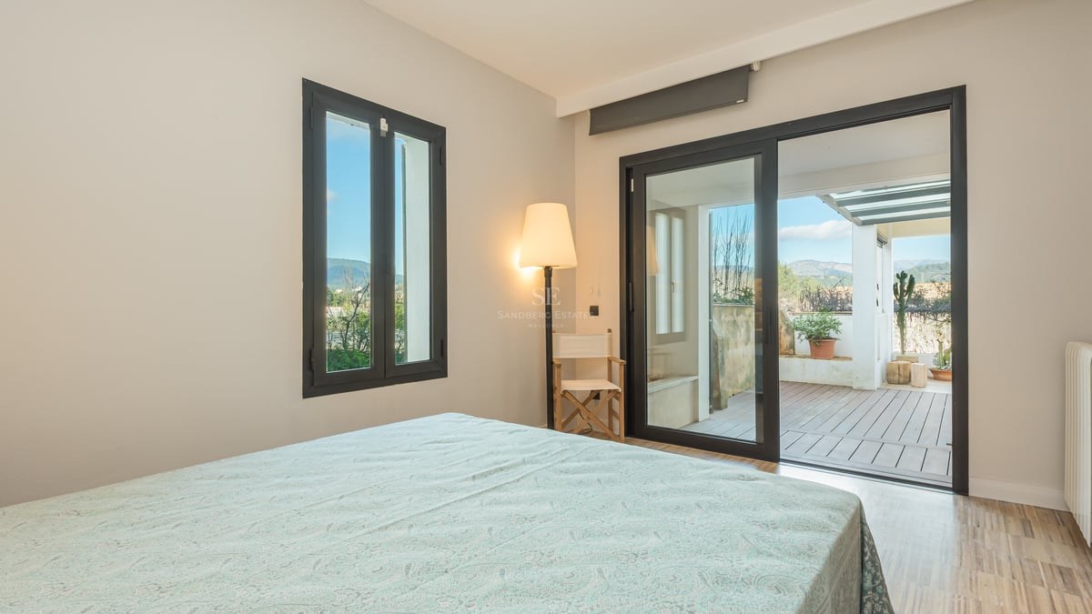 Ljust sovrum med skjutdörrar i glas som leder ut till en träterrass med bergsutsikt.