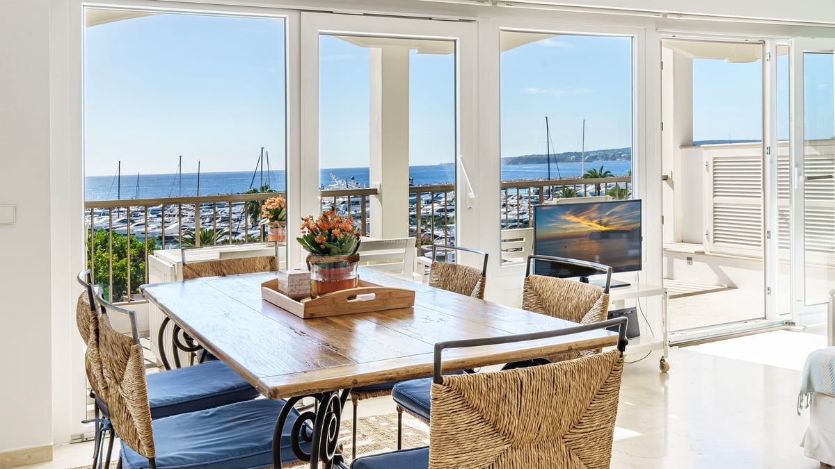 Sunlit dining room with a wooden table and woven chairs looking out over a yacht-filled marina and the blue ocean.