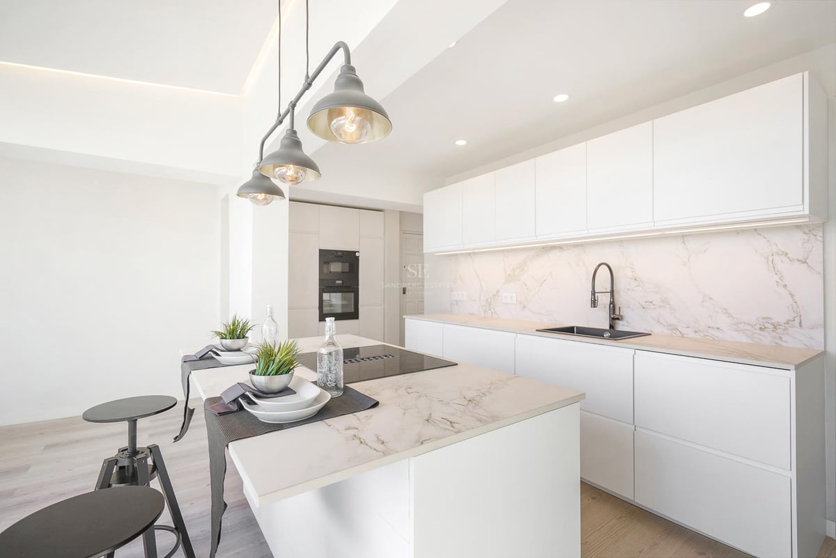 Moderne weiße Küche mit großer Marmorinsel, integrierten Geräten und industriellen Pendelleuchten.