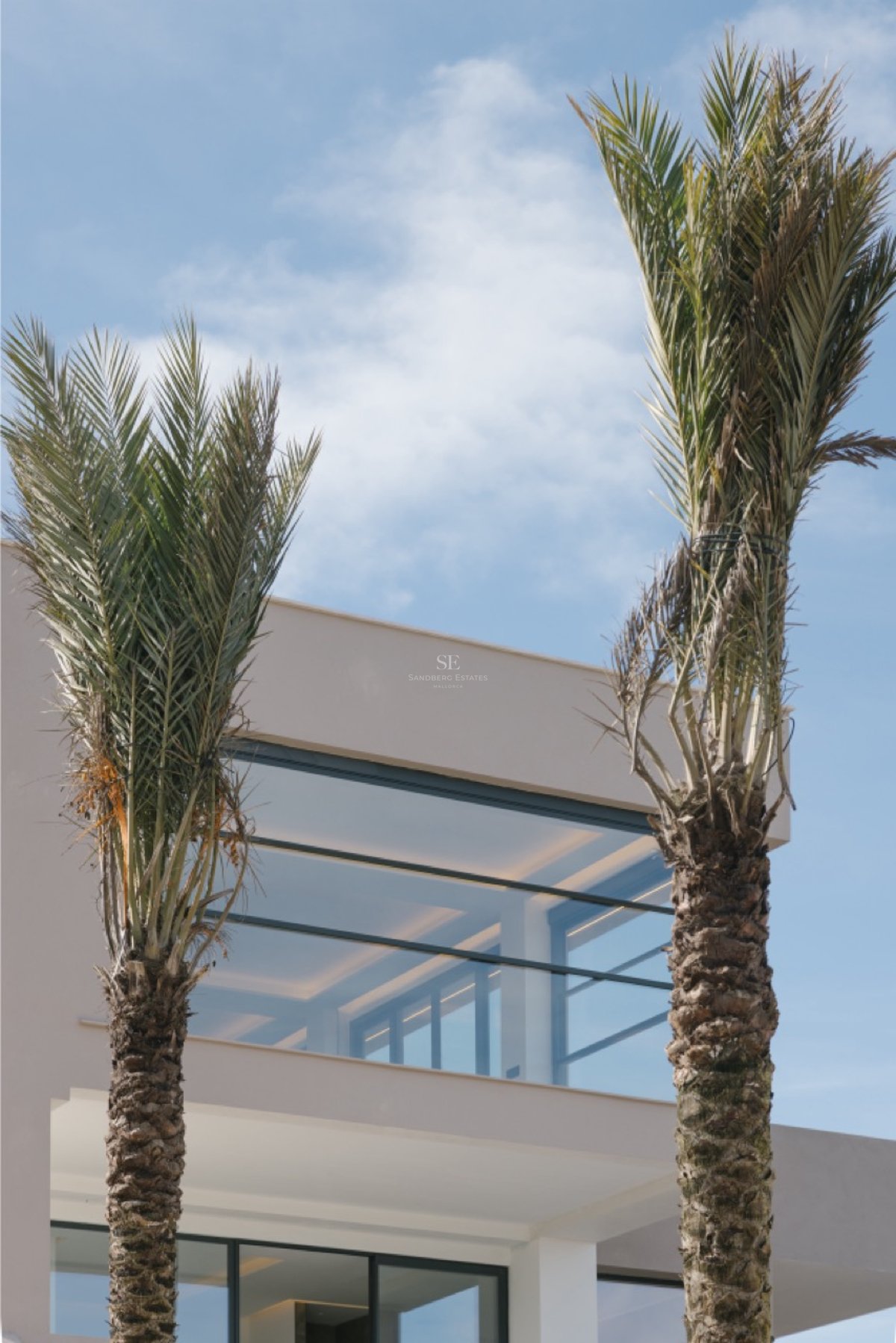 Außenansicht einer modernen weißen Villa mit bodentiefen Glasfronten und Palmen vor blauem Himmel.