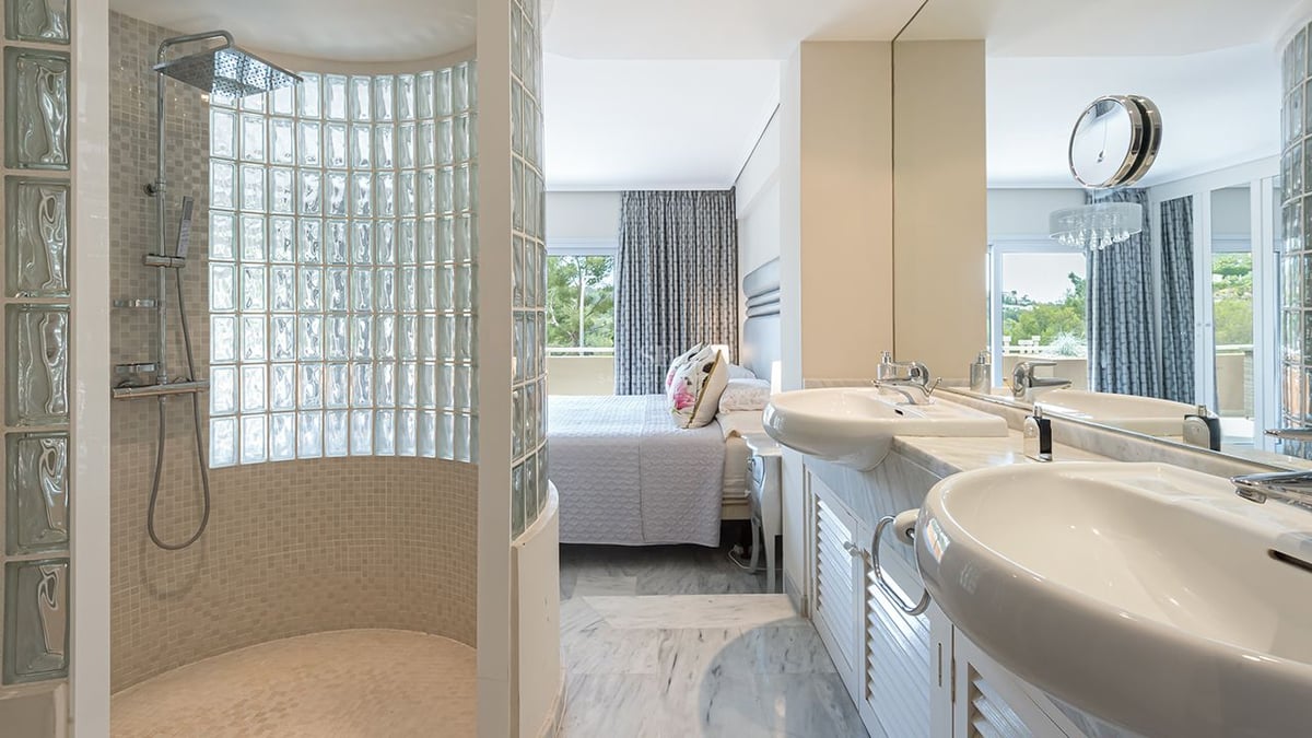 En-suite bathroom featuring double marble vanity and glass block shower, adjacent to a bright bedroom area.
