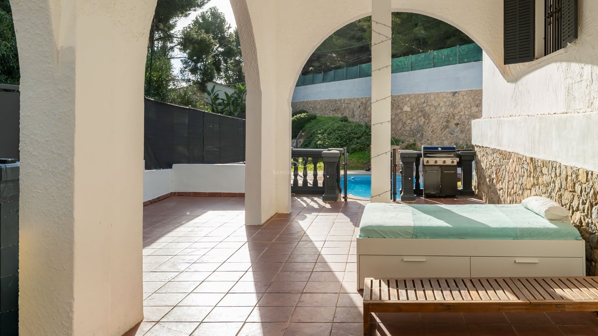 Terrasse couverte avec arches blanches, lit de repos, barbecue et vue sur une piscine ensoleillée.