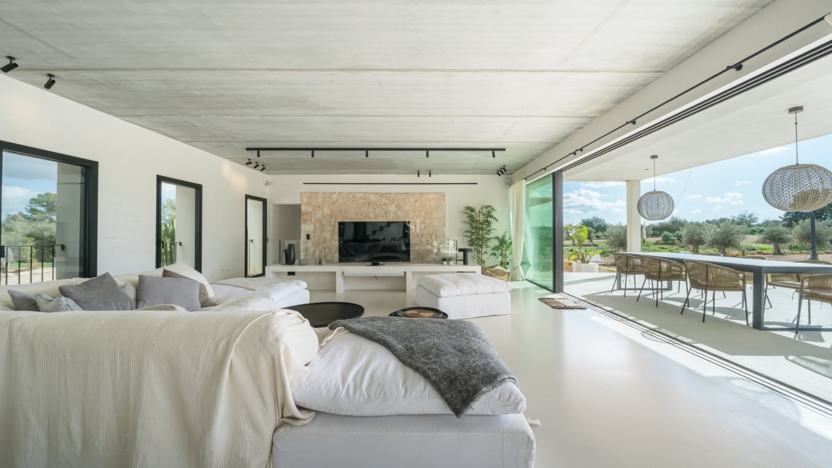 Modernt vardagsrum med vita soffor, betongtak, stenvägg och skjutdörrar i glas som leder ut till en terrass.