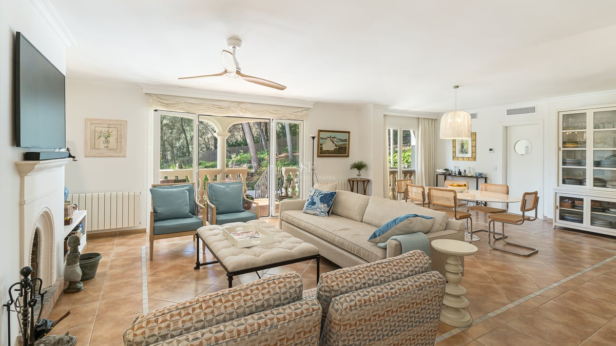Spacious living room with terracotta floors, cream sofas, fireplace, and glass doors opening to a lush garden.