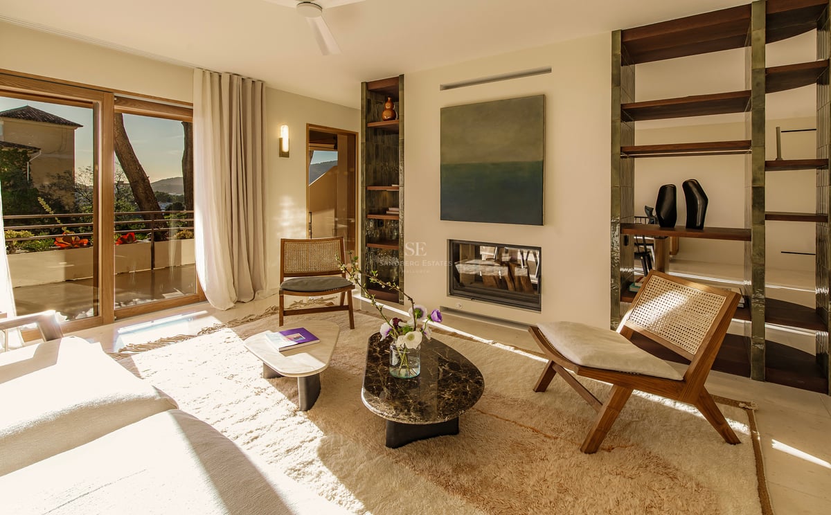 Helles Wohnzimmer mit Rattanstühlen, Marmortischen, Einbaukamin und großen Glastüren mit Blick ins Freie.