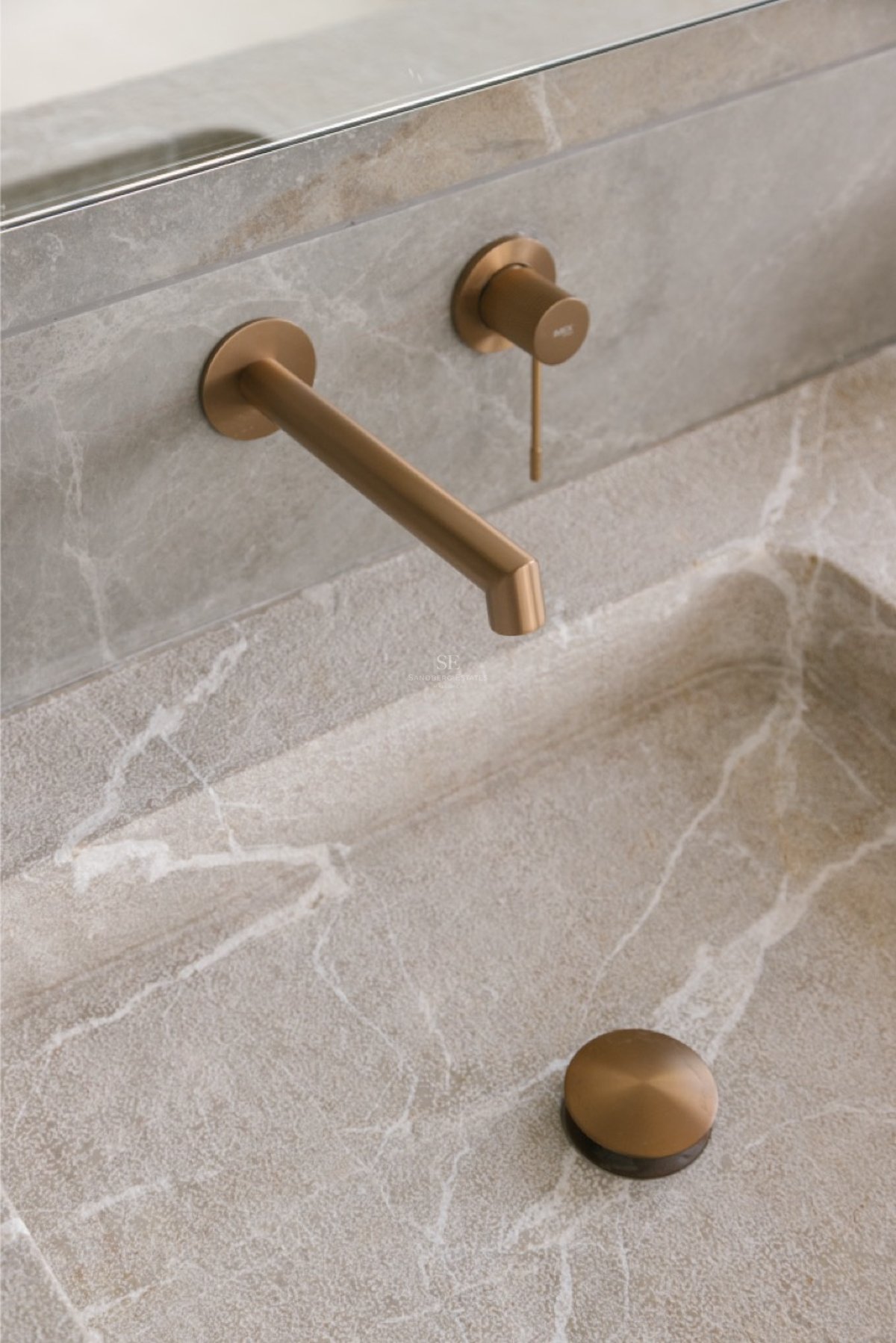 Wandmontierte Armatur aus gebürsteter Bronze über einem Waschbecken aus Naturstein mit weißer Maserung.