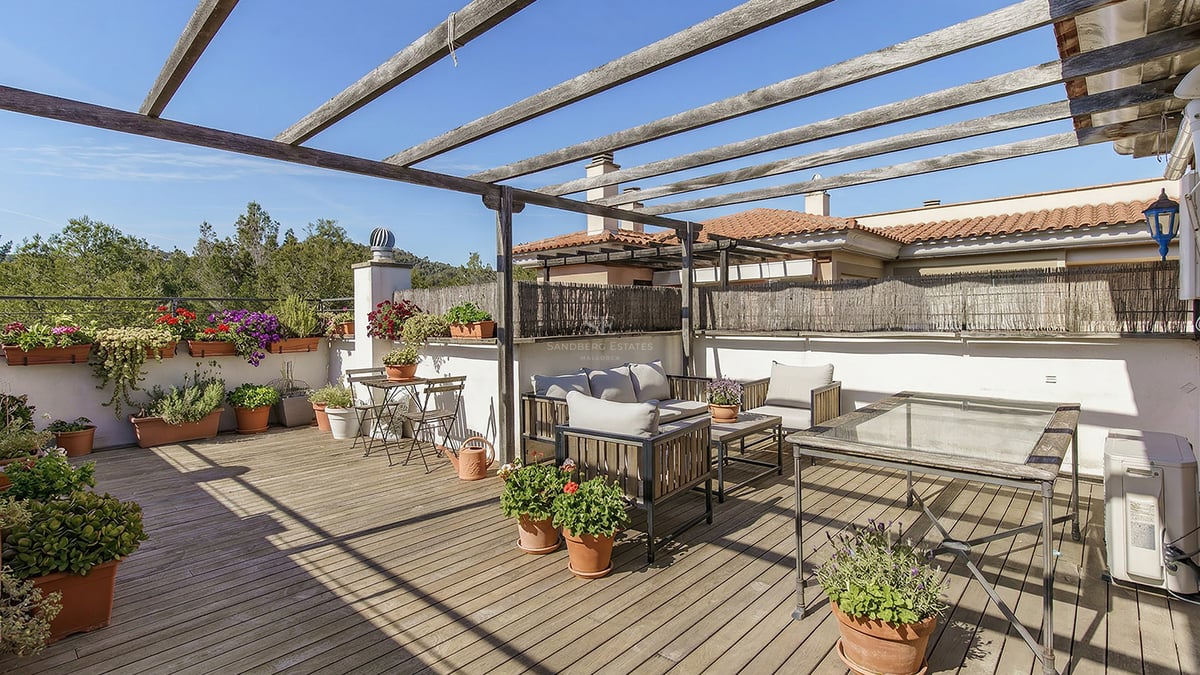 Takterrass med träpergola, trädäck, olika krukväxter och utemöbler under en klarblå himmel.