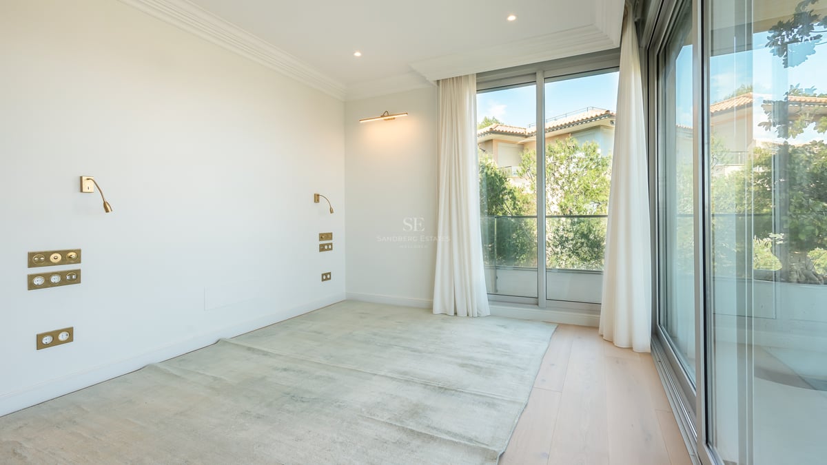 Empty modern bedroom with light wood floors, floor-to-ceiling glass doors, white walls, and elegant brass wall lamps.