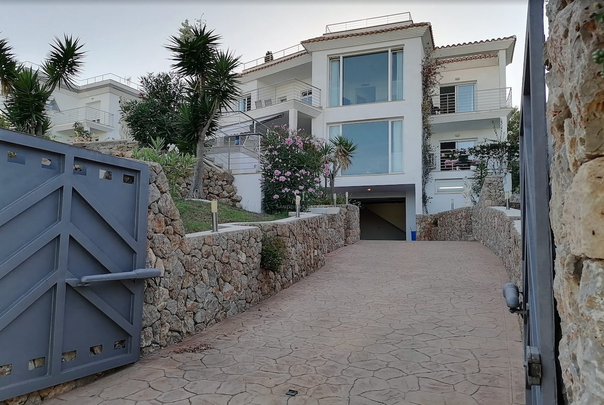 Villa blanche à plusieurs niveaux avec grandes baies vitrées, vue depuis une allée sécurisée avec murs en pierre.