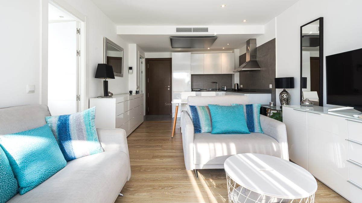 Salon lumineux avec canapés blancs, coussins turquoise, parquet en bois et cuisine moderne en arrière-plan.