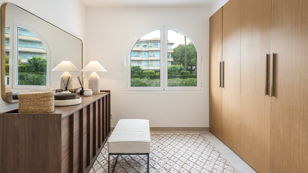 A modern dressing room featuring large oak wardrobes, an arched window, a mirrored console, and a cushioned bench on a rug.