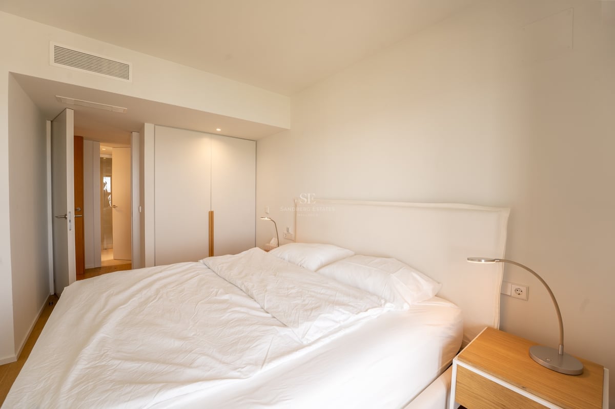 Ljust sovrum med stor säng, vita sängkläder, inbyggd garderob och sängbord i trä.