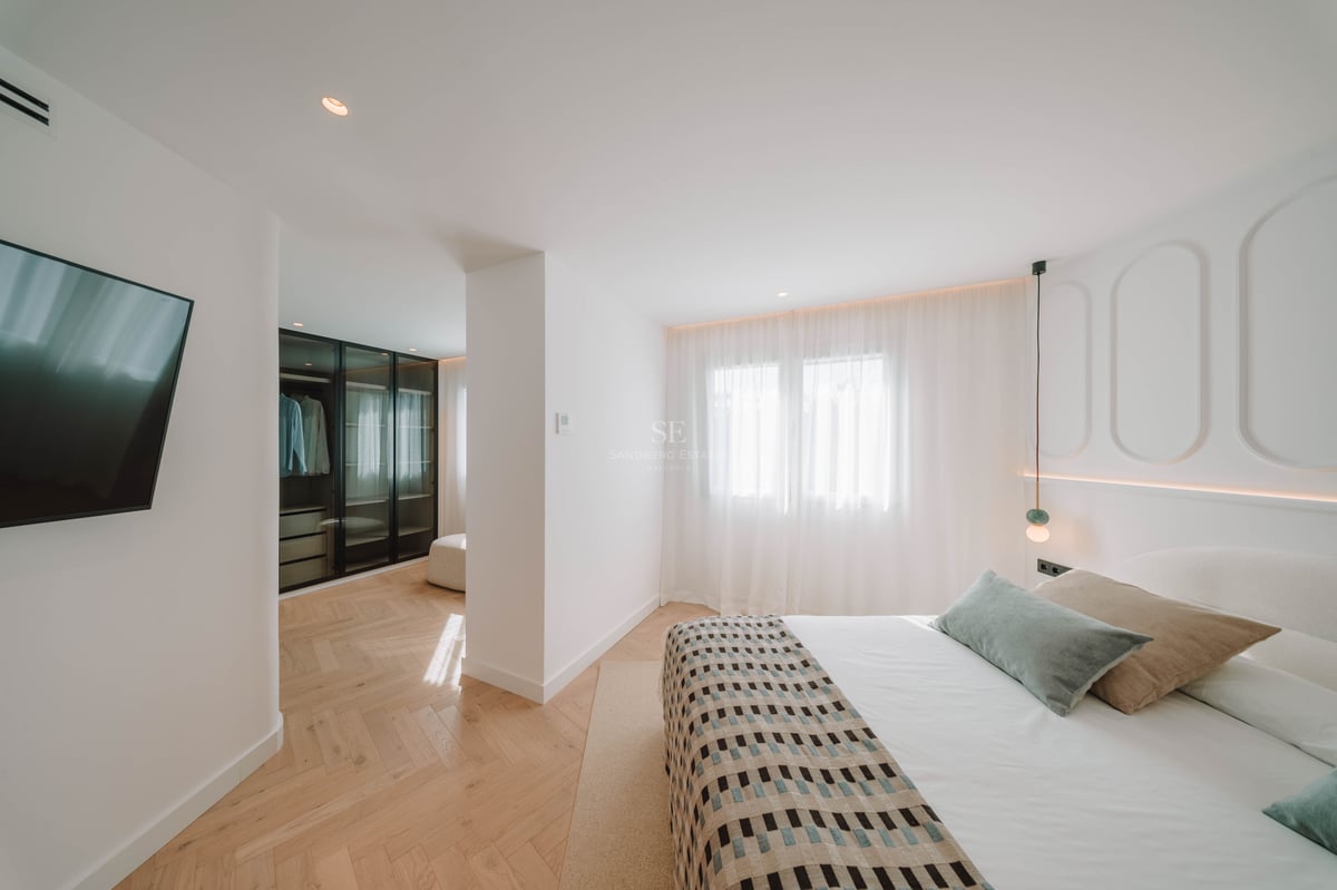 Chambre principale lumineuse avec parquet en chevrons, grand lit et dressing vitré en arrière-plan.