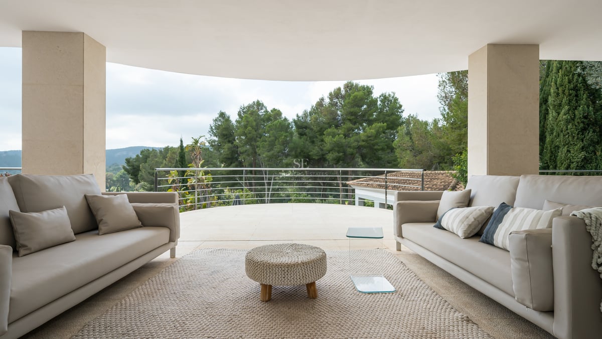 Terrasse moderne avec canapés gris et pouf tressé donnant sur une forêt dense de pins.