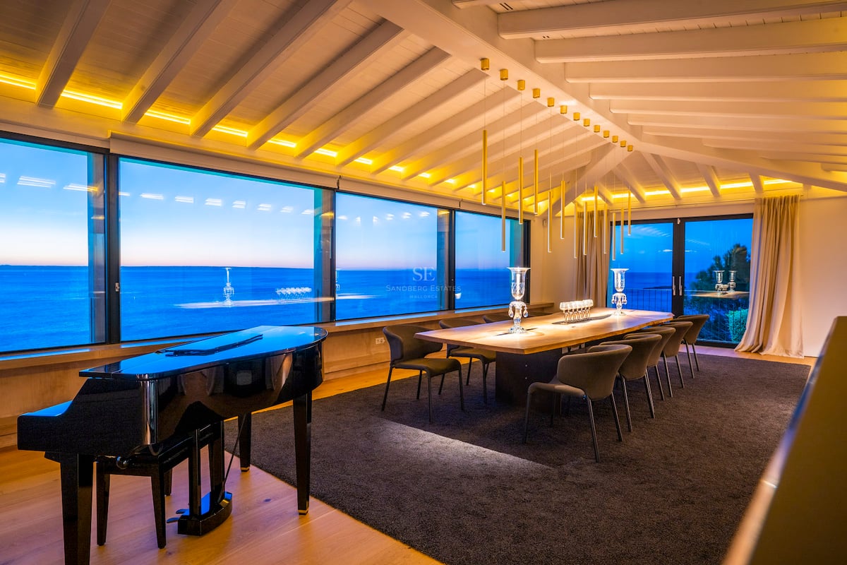 Ein modernes Esszimmer mit Flügel, großem Holztisch und bodentiefen Fenstern mit Blick auf das Meer in der Dämmerung.