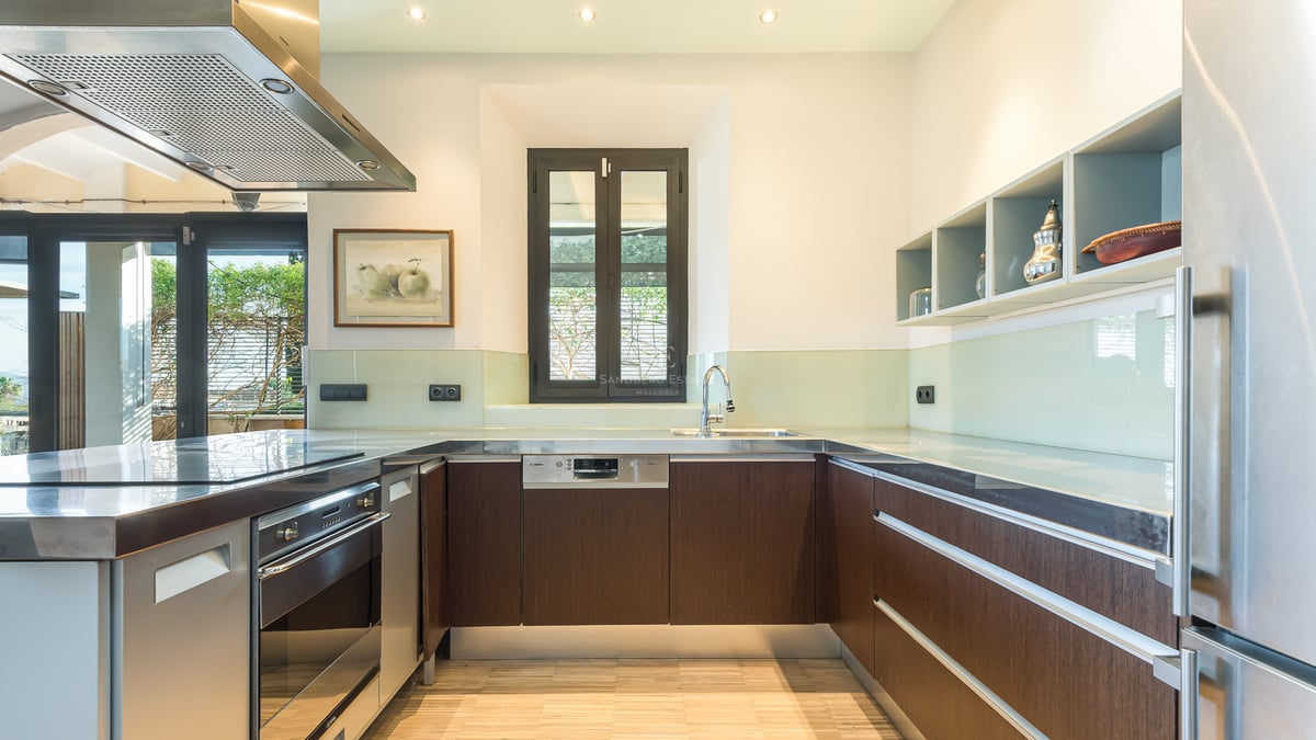 Modernt kök med mörka träskåp, bänkskivor i rostfritt stål och stänkskydd i glas under ljus belysning.