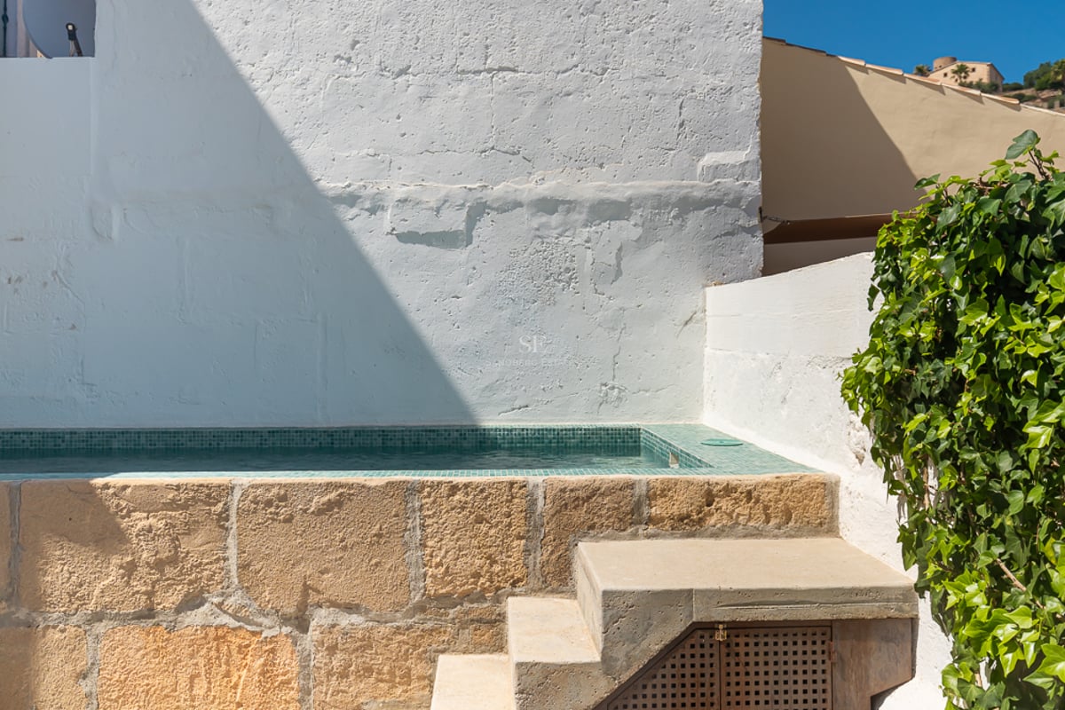 Petite piscine surélevée avec mosaïque turquoise et socle en pierre naturelle contre un mur blanc et du lierre.