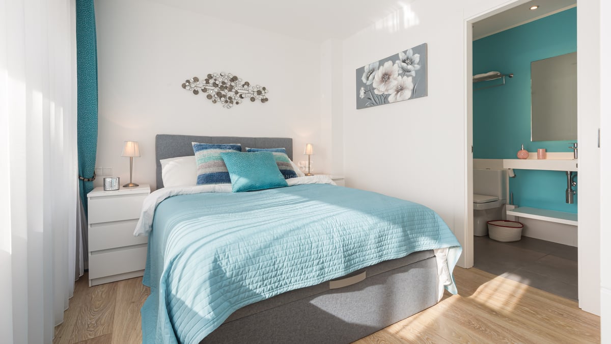 Chambre lumineuse avec lit turquoise, tête de lit grise et salle de bain privative visible en arrière-plan.