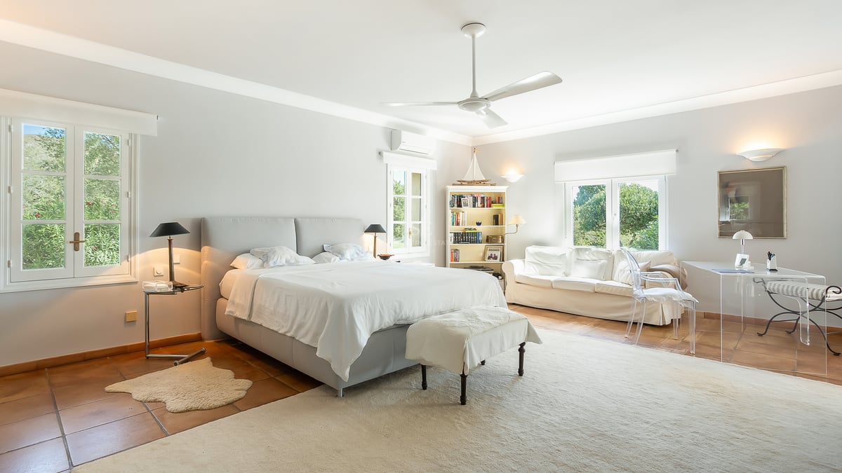 Grande chambre parentale avec sol en terre cuite, canapé blanc, bureau en verre et fenêtres ouvrant sur le jardin.