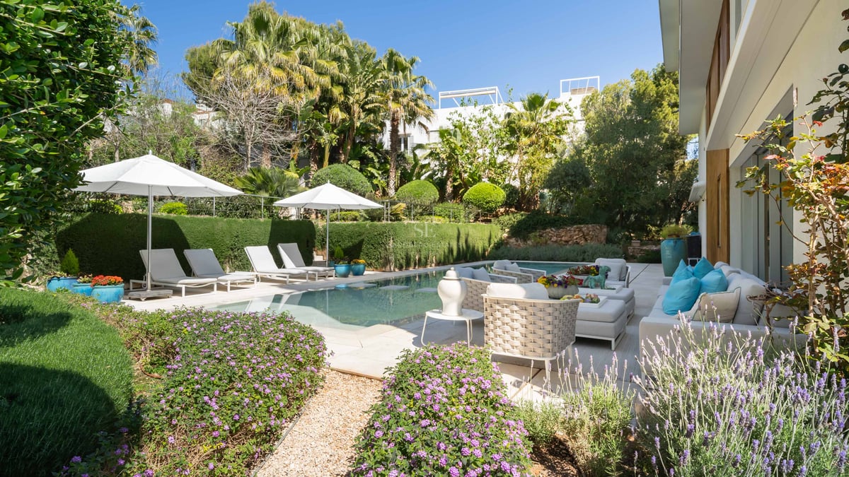 Ein heller Swimmingpool umgeben von üppigem Grün, weißen Sonnenschirmen und hochwertigen Loungemöbeln.