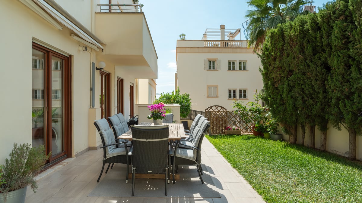 Espace repas extérieur sur terrasse carrelée bordant un jardin avec pelouse et haies hautes sous le soleil.