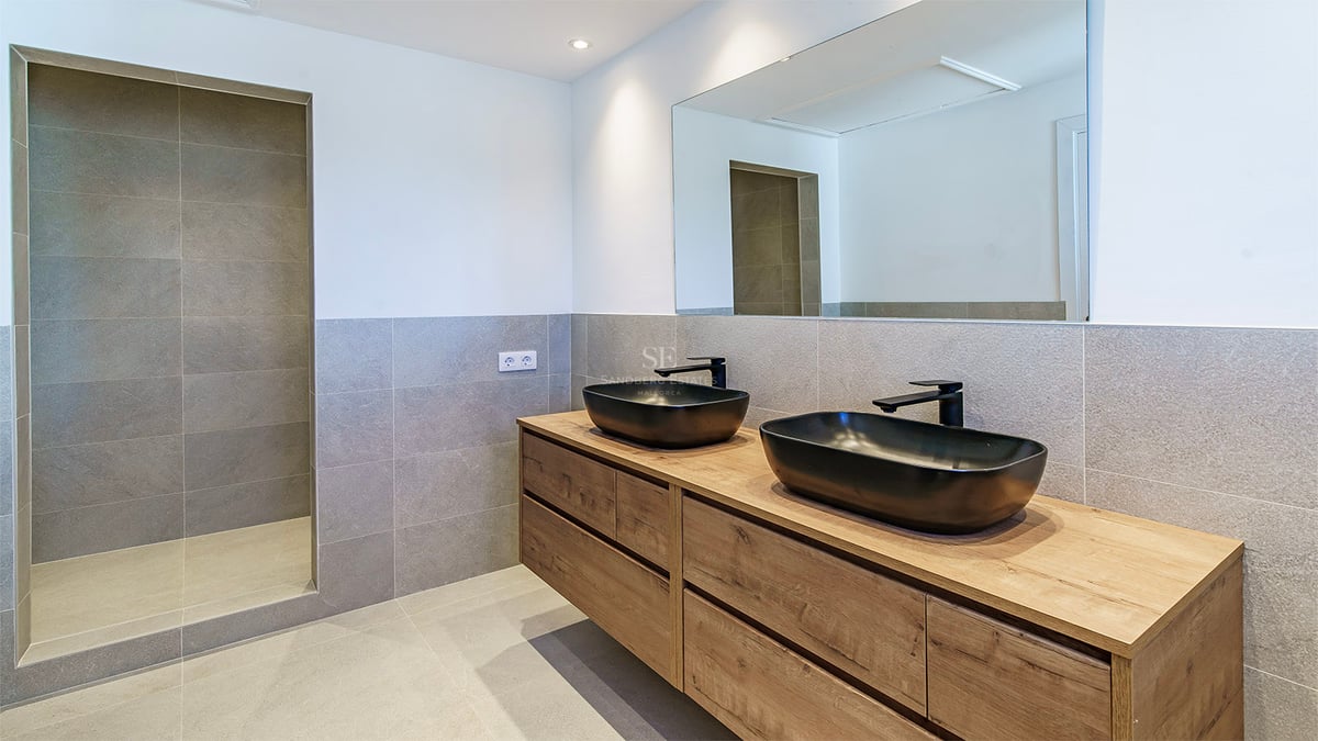 Samtida badrum med grått kakel, dubbelkommod i trä, svarta tvättställ och en rymlig duschkabin.