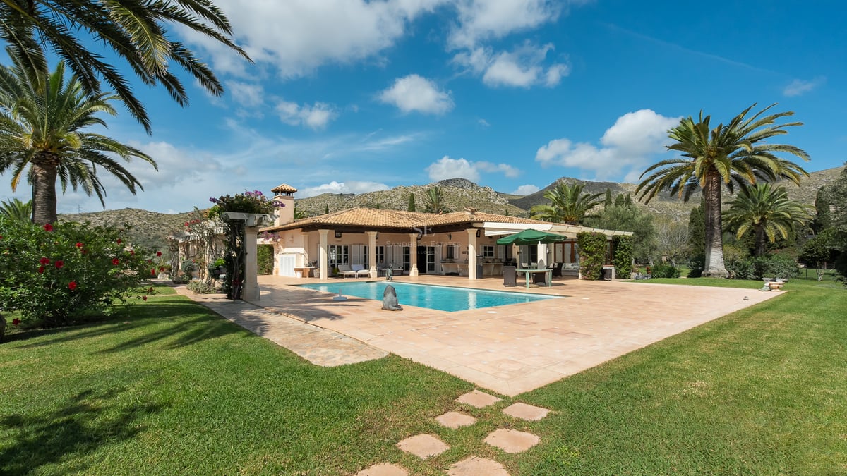 Villa espagnole avec piscine rectangulaire, terrasse en terre cuite, palmiers et montagnes sous un ciel bleu.