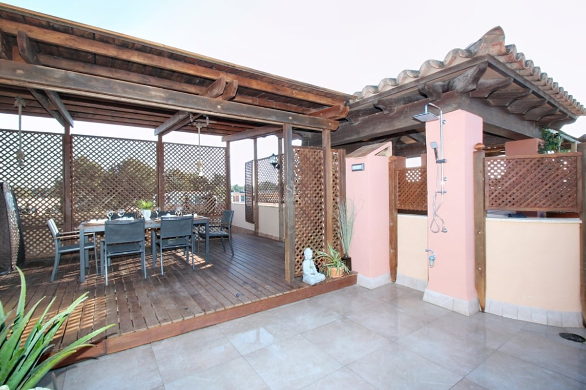 En träpergola över en matgrupp på ett mörkt trädäck, med en utedusch monterad på en rosa vägg.