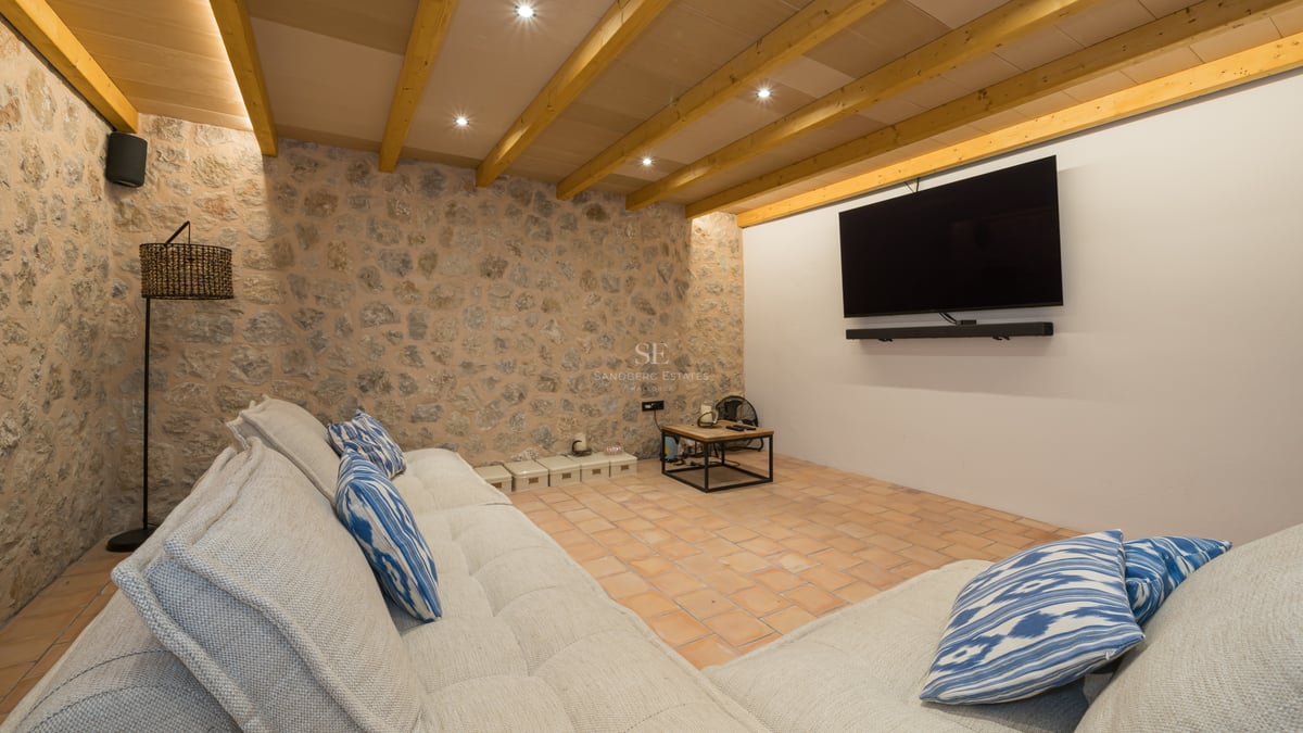 Salon multimédia rustique avec un grand canapé, télévision murale, murs en pierre et poutres apparentes.