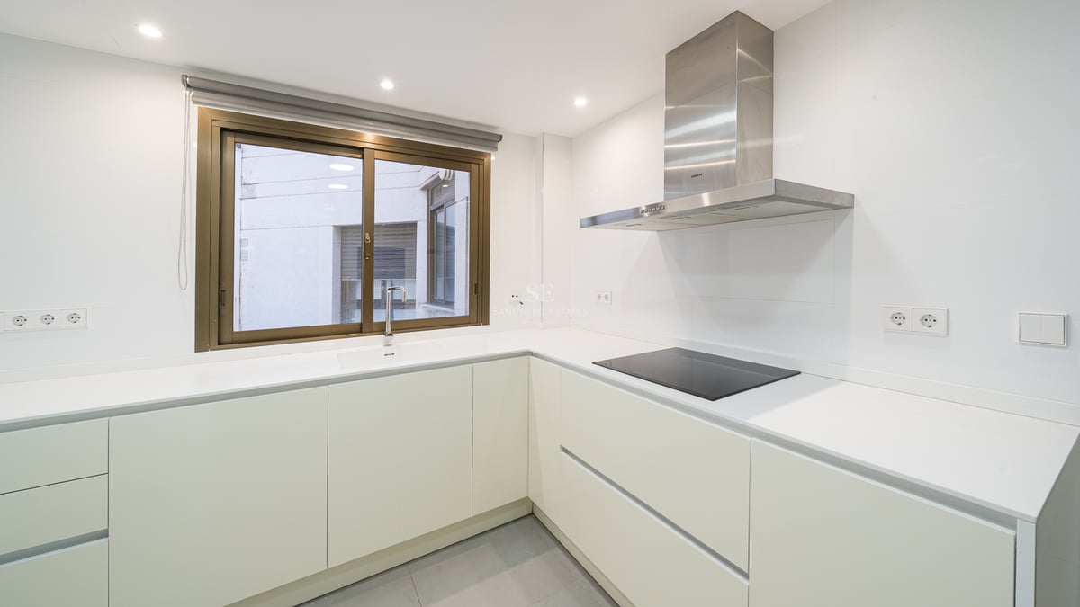 Minimalistiskt vitt kök med L-formad bänkskiva, induktionshäll och köksfläkt i rostfritt stål.