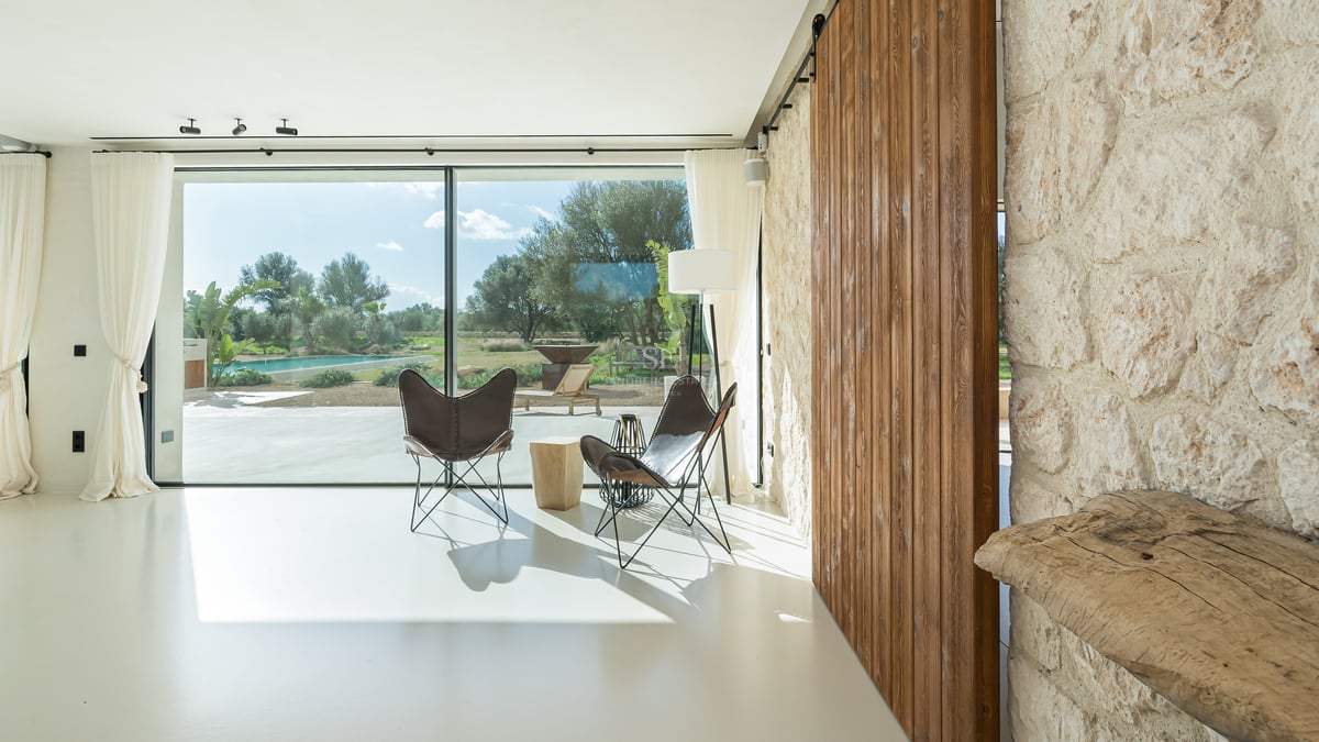 Ljust vardagsrum med stora glaspartier, butterfly-stolar, skjutdörr i trä och vägg i natursten.