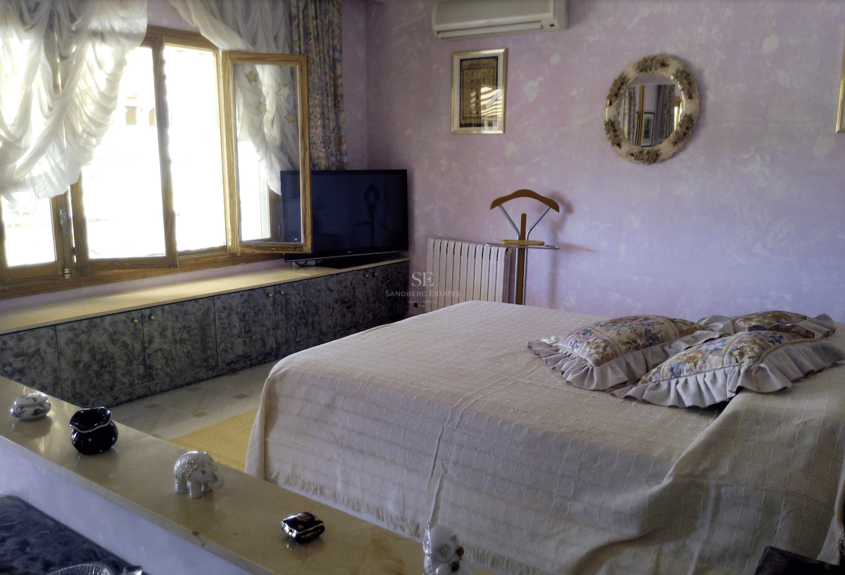 Dormitorio con paredes de color lila, cama grande con cojines florales y amplios ventanales de madera bajo luz natural.