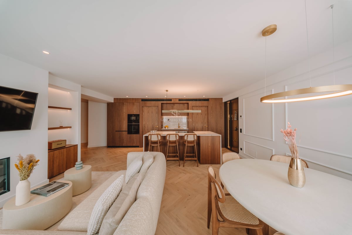 Grand salon luxueux avec canapé beige, parquet en chevrons et cuisine intégrée en bois naturel.