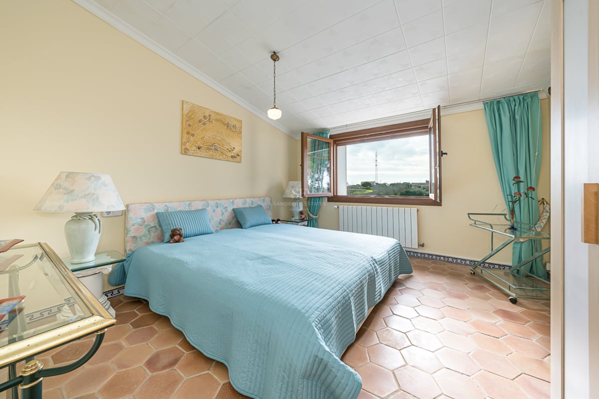 Chambre avec sol en terre cuite, lit bleu et fenêtre ouverte donnant sur un paysage verdoyant.