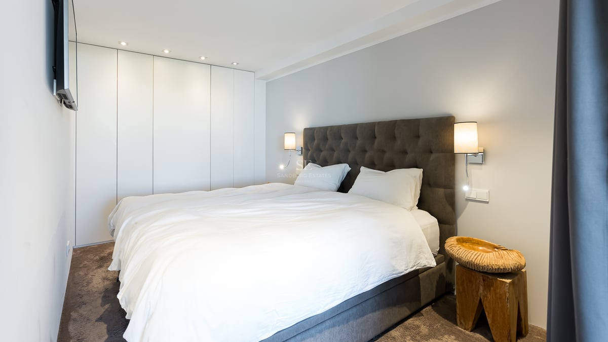 Zeitgenössisches Schlafzimmer mit weißem Bett, grauem Polsterkopfteil, weißen Wandschränken und Designer-Holzhocker.