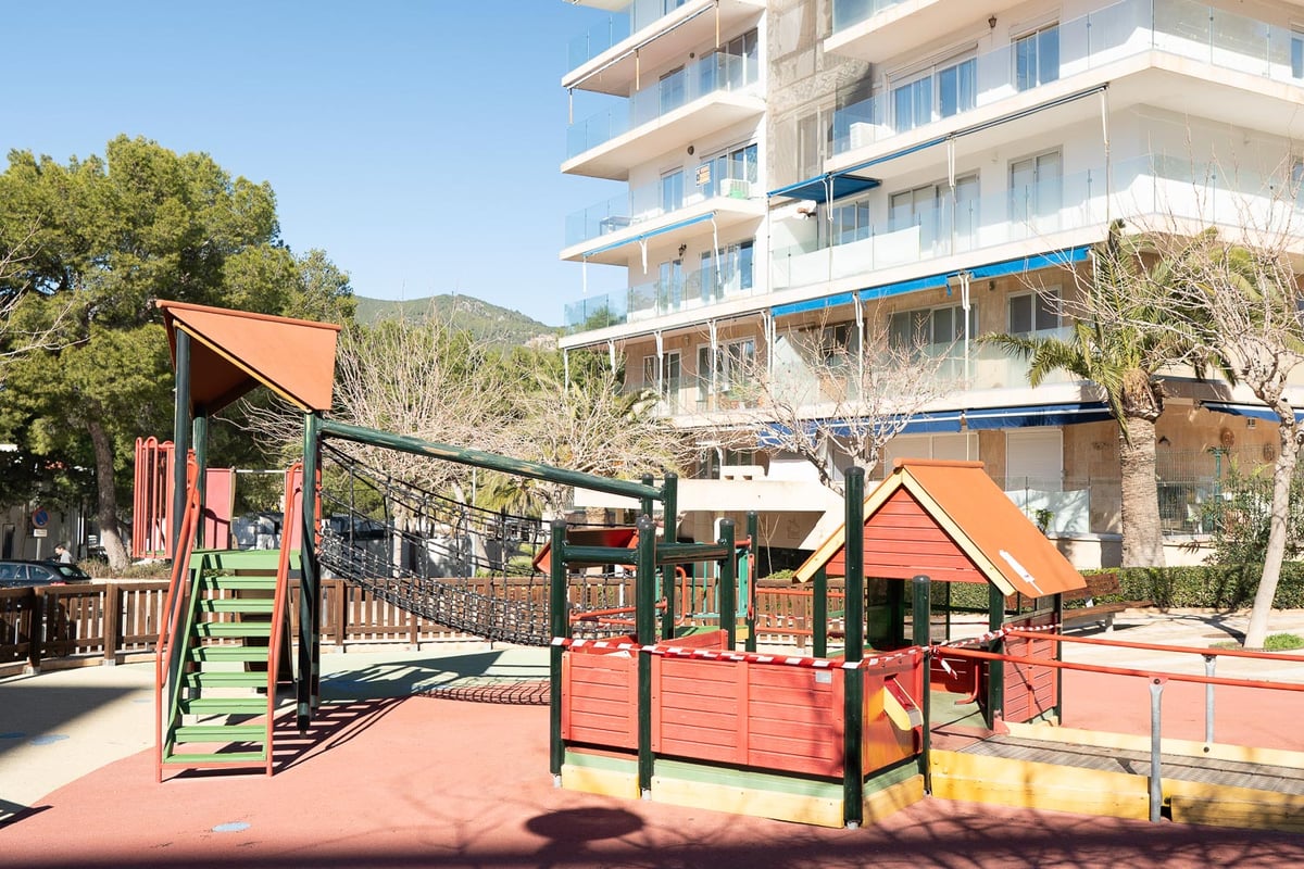 Kinderspielplatz mit Holzgeräten und Sicherheitsboden vor einem modernen Apartmenthaus.