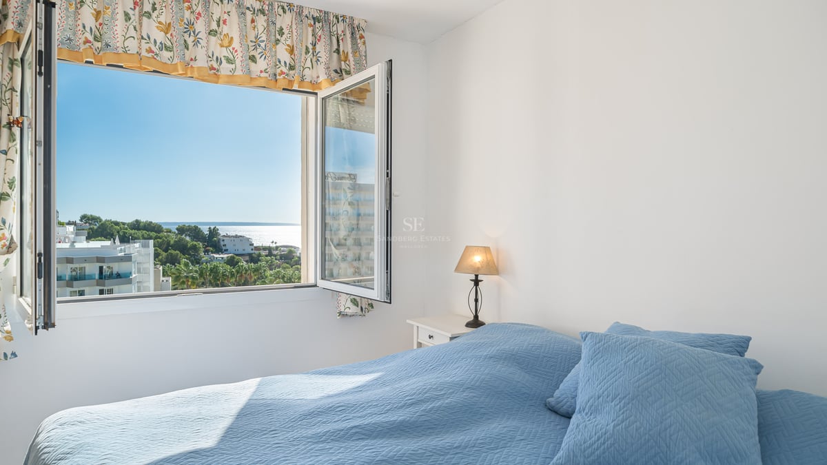A sunlit bedroom featuring a blue quilted bed and an open window looking out over trees and the sparkling sea.