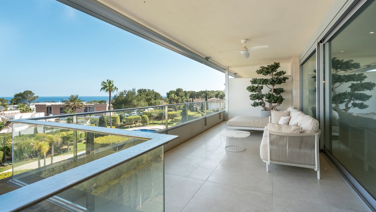 Wide modern terrace with white outdoor sofas, glass railings, and views of lush gardens and the sea in the distance.