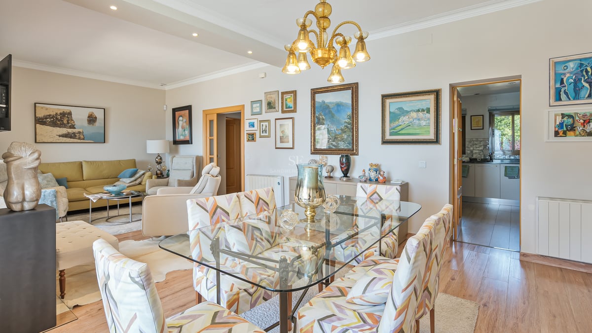 Ljust rum med öppen planlösning, matbord i glas, mönstrade stolar och konstsamling på väggen.