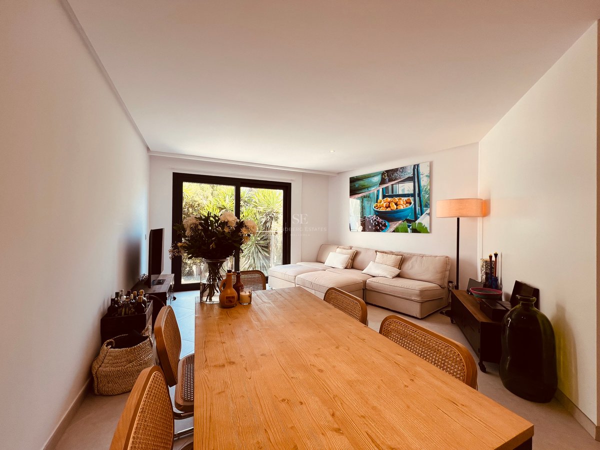 Salon et salle à manger avec grande table en bois, chaises en rotin et canapé beige près d'une baie vitrée.