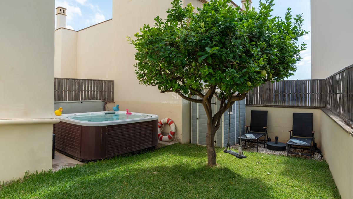 Petit jardin privé avec pelouse, jacuzzi en bois, un citronnier central et deux chaises longues noires.