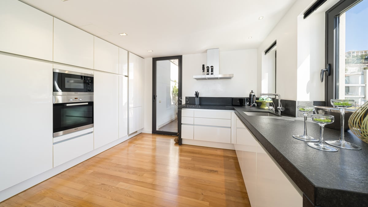 Modern white kitchen with dark stone countertops, oak flooring, integrated appliances, and access to a terrace.