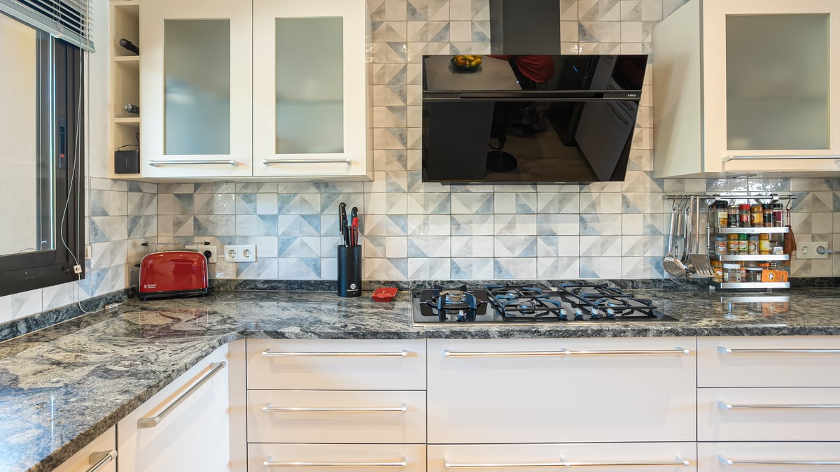 Närbild av ett modernt kök med bänkskiva i granit, gasspis, mönstrat kakel och vita skåpluckor.