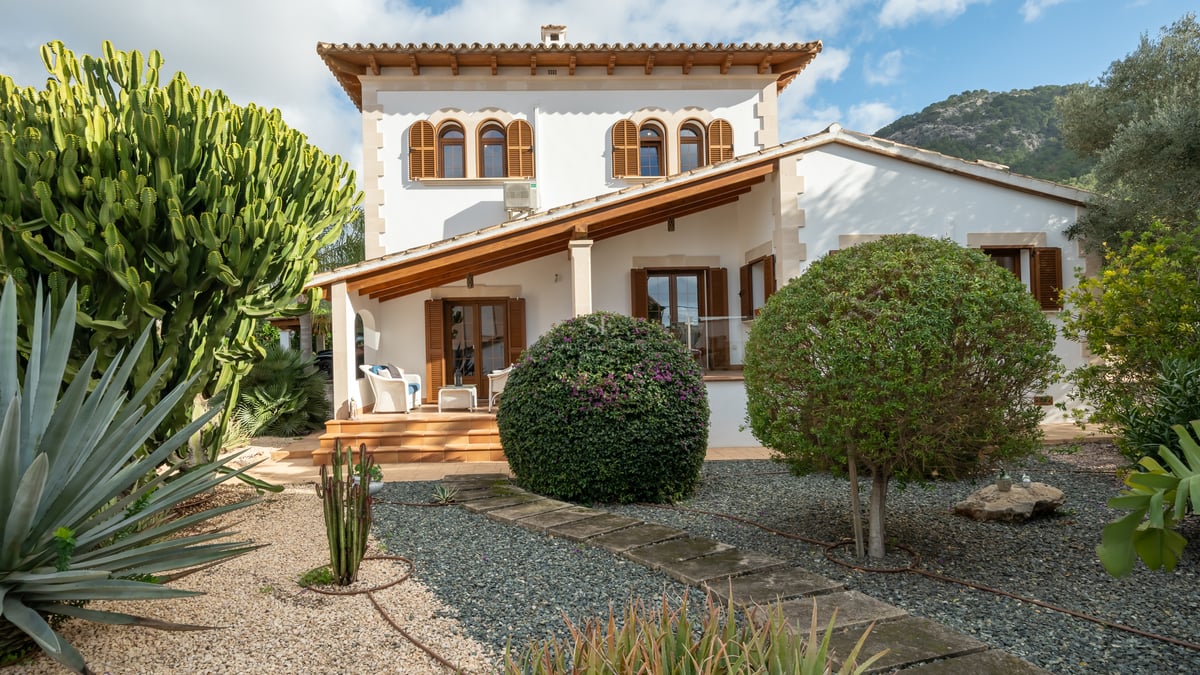 White villa facade featuring wooden shutters, terracotta roof, and a gravel garden with cacti and agave plants.