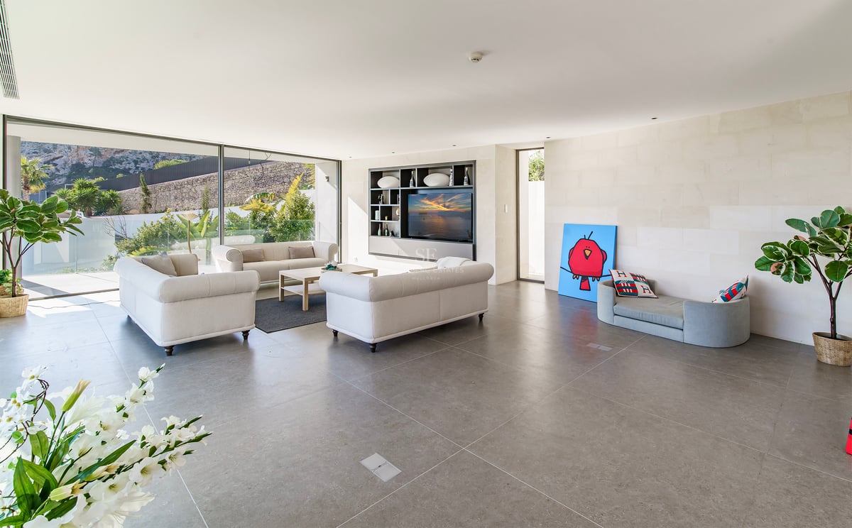 Salon moderne et lumineux avec canapés beiges, sol en pierre grise et grandes baies vitrées donnant sur un jardin.