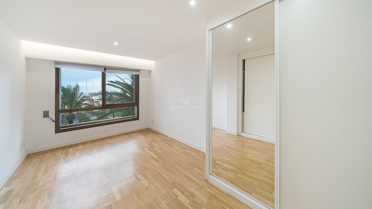 Tomt sovrum med ljust trägolv, spegelgarderob och fönster med utsikt över palmer och båtar.