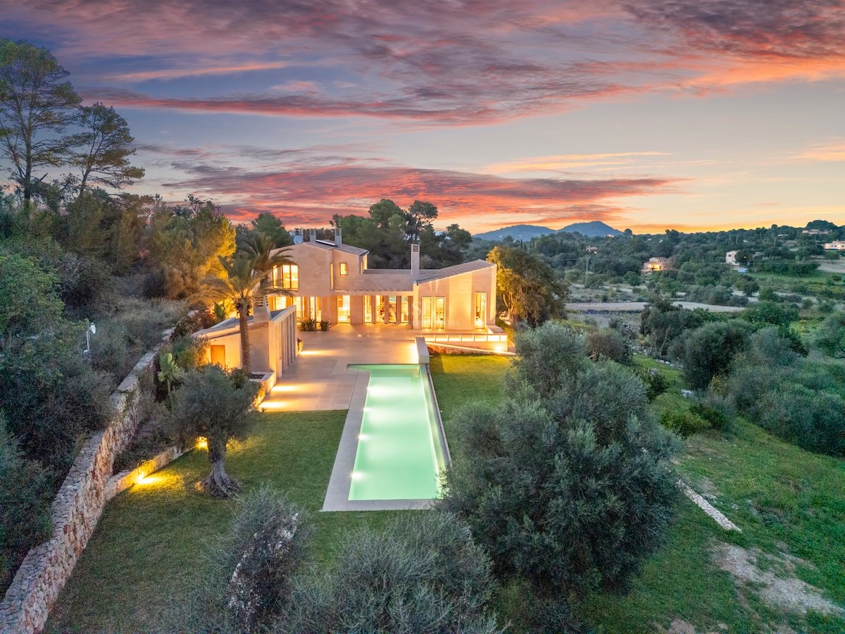Vue aérienne d'une villa moderne avec piscine lumineuse, murs en pierre et ciel de coucher de soleil coloré.